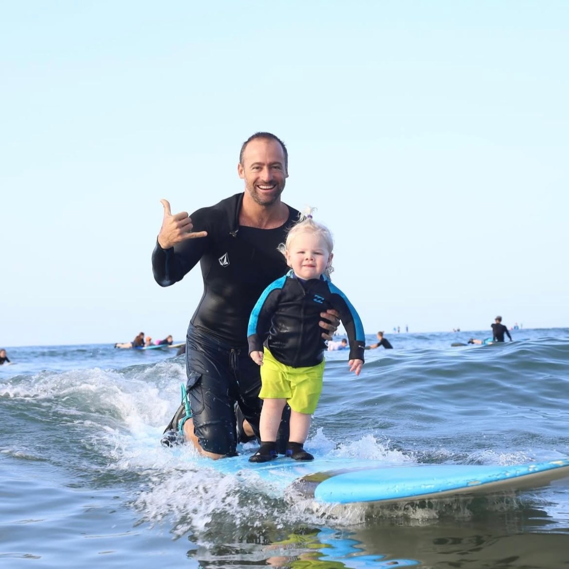 Man and child surfing together, man doing a shaka sign, other surfers in background.