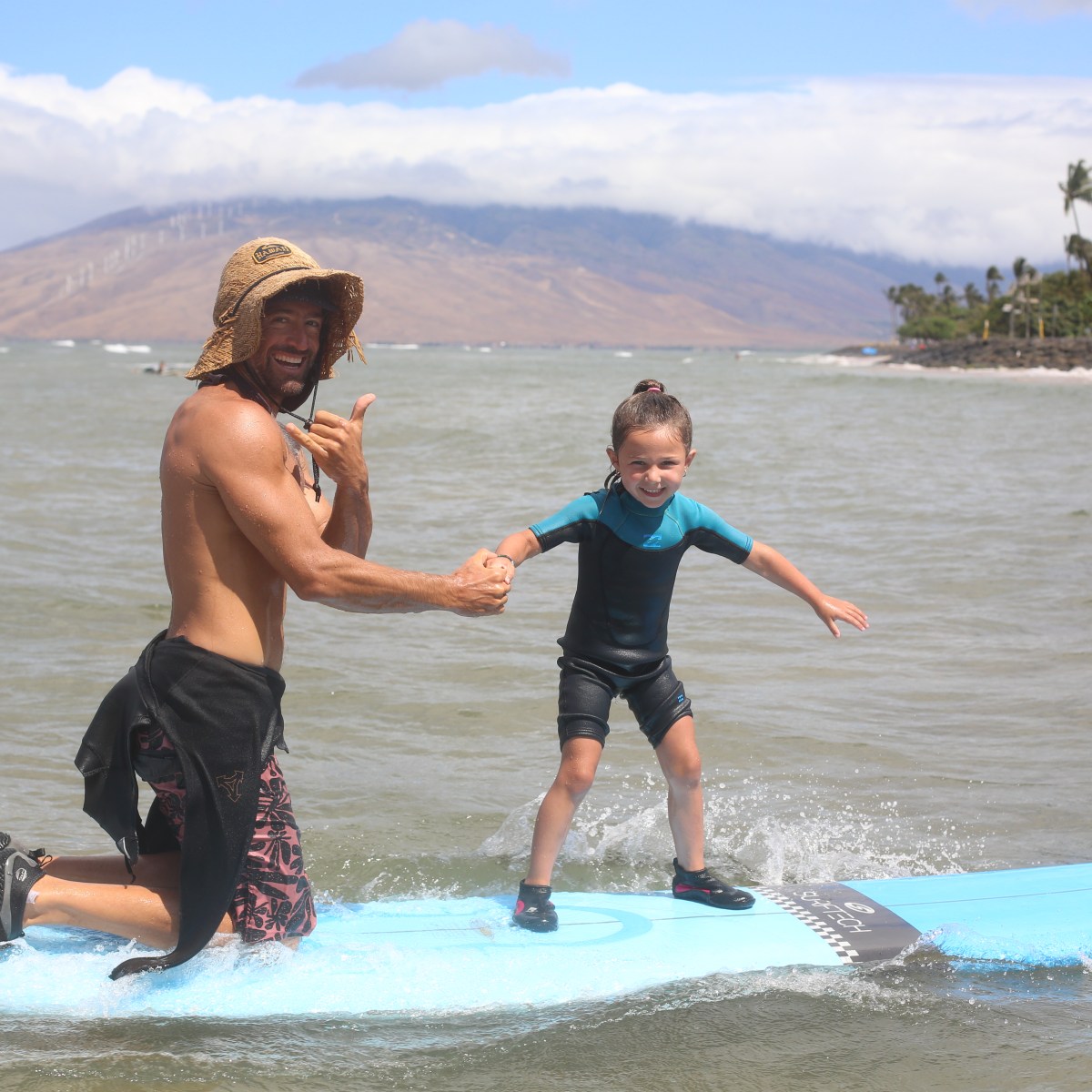 Man and child surfing on a blue board with mountains in the background.