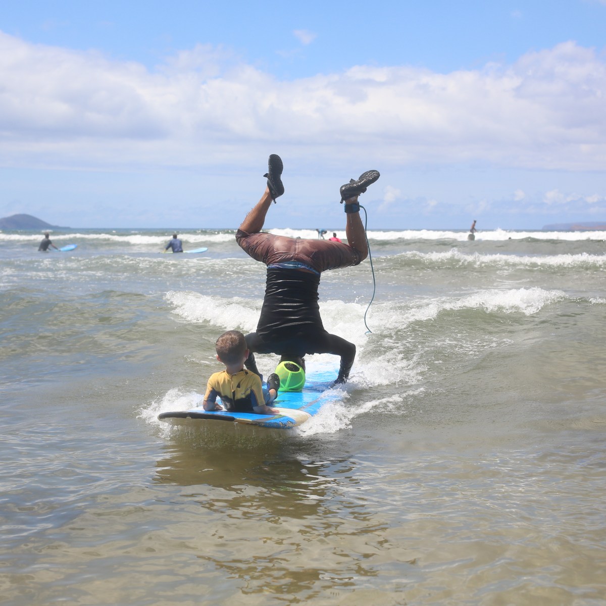 Man doing headstand on surfboard with child sitting in front in shallow ocean water.