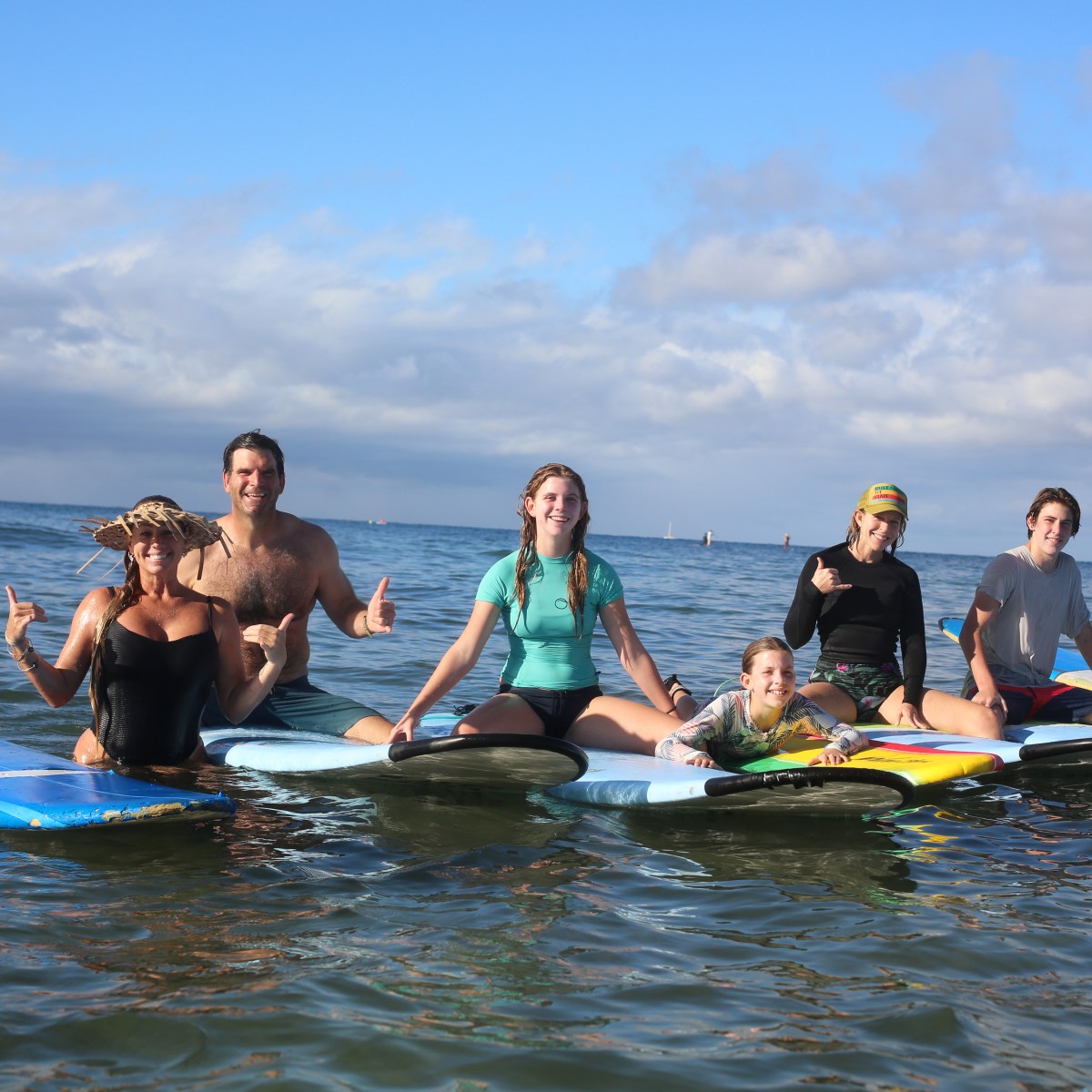 Six people sitting on surfboards in the ocean, smiling under a partly cloudy sky.