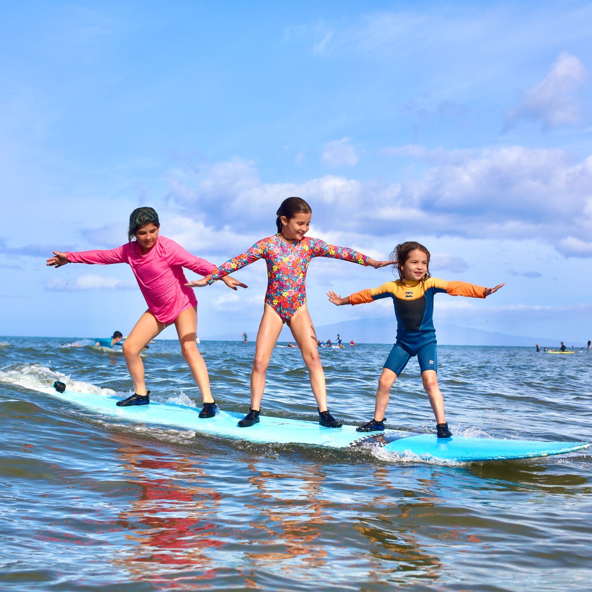 Three children balancing on a surfboard in the ocean under a blue sky.