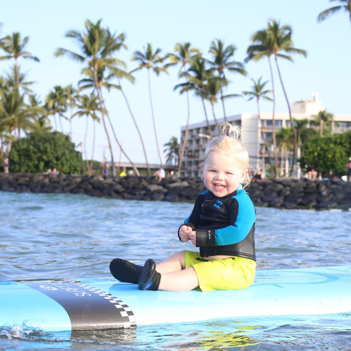 Smiling toddler sits on a surfboard in the ocean, with palm trees in the background.
