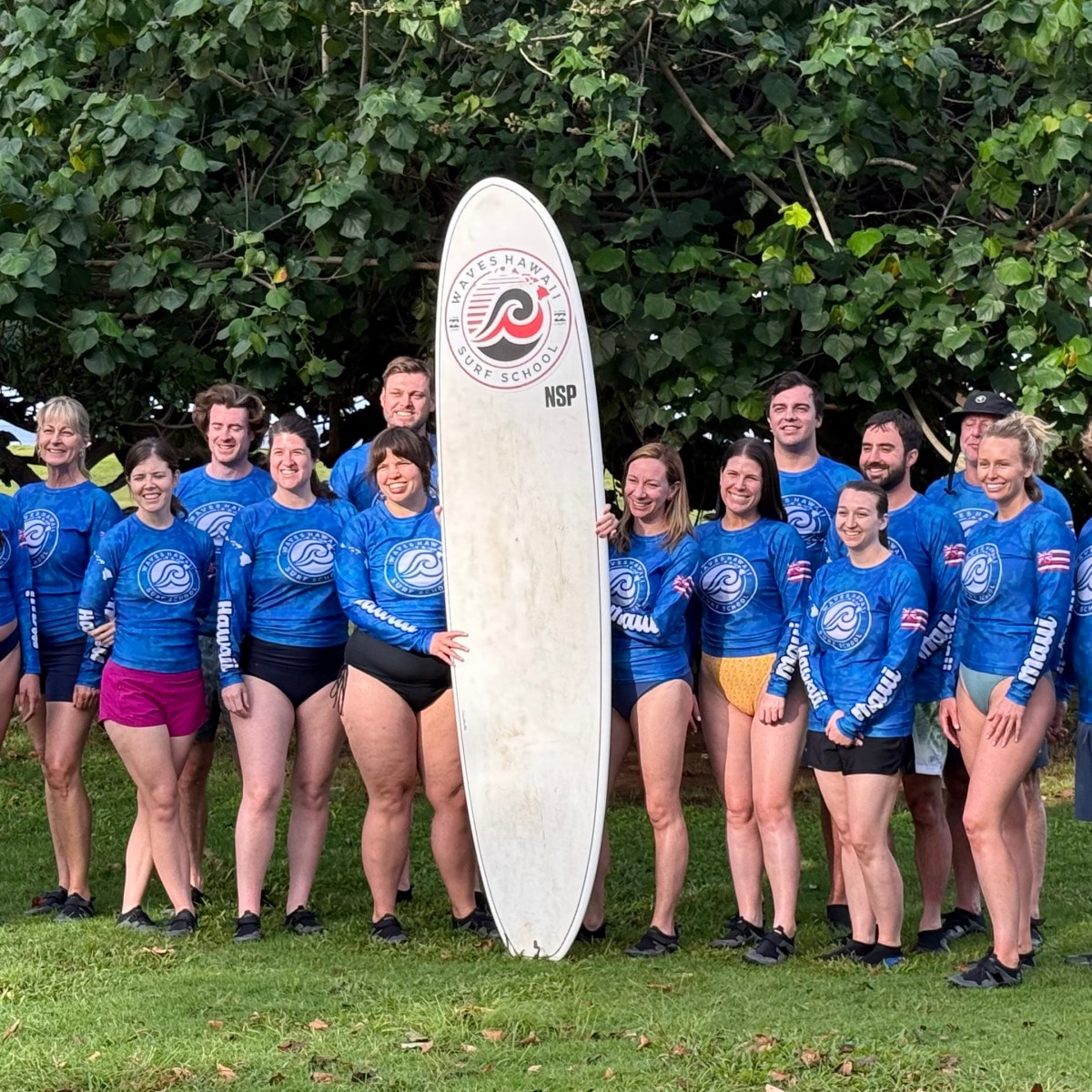 Group of people in blue shirts with a surfboard, standing on grass in front of trees.
