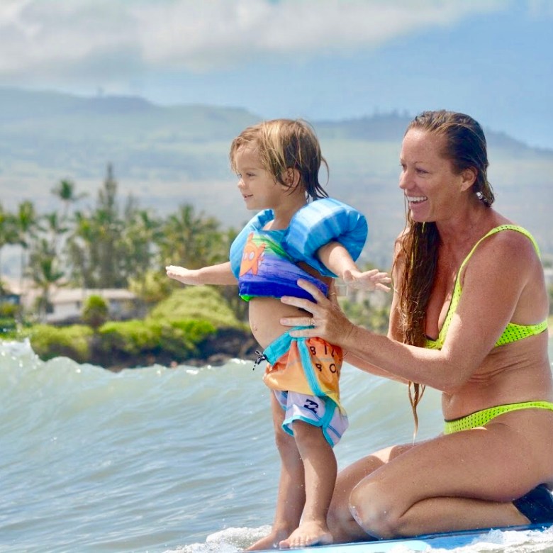 Smiling woman holding child on surfboard in ocean with beach in background.