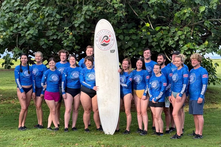 Group in blue shirts posing with surfboard in front of a large tree.