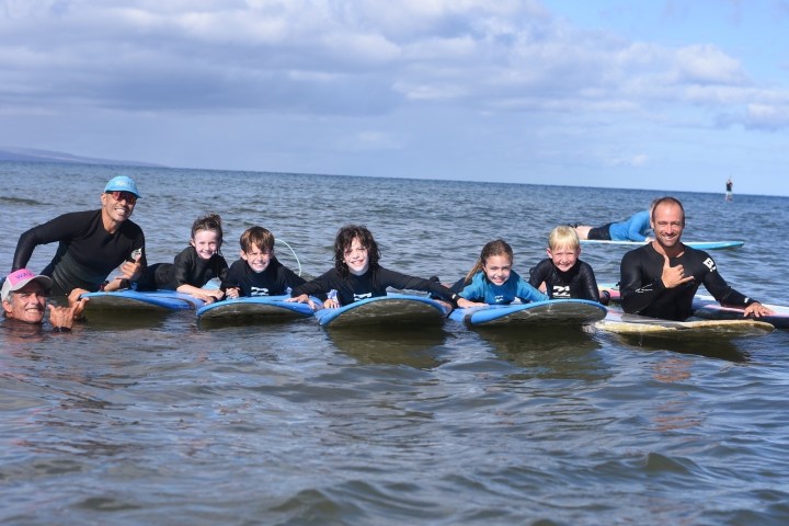 Group of kids and adults on surfboards in the ocean, smiling.