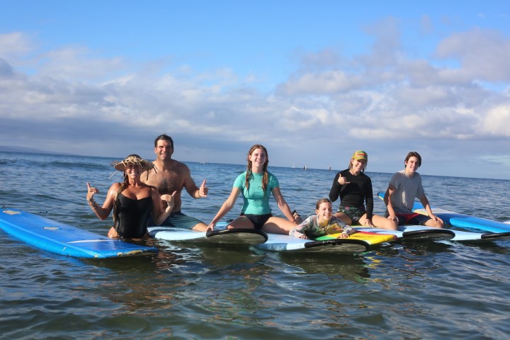 Group of six people sitting on surfboards in calm ocean water, smiling and gesturing thumbs up.