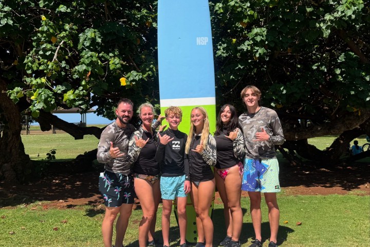 Six people posing with surfboards in front of trees on a sunny day.