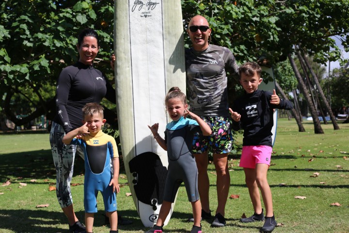 Family in wetsuits posing with surfboard in park.